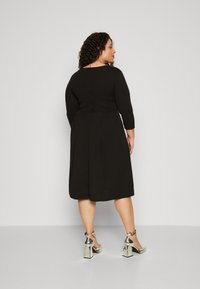 Vestido negro hasta la rodilla con mangas de tres cuartos y cremallera trasera. Combinado con zapatos de tacón bloque metálicos en plata, que muestran correas en los tobillos.