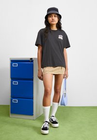 Black T-shirt with graphic, tan corduroy shorts, white knee-high socks, black sneakers, and a striped bucket hat. Light blue handbag.