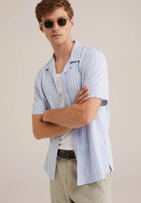 Chemise à manches courtes, rayée bleu clair, fabriquée en tissu avec une coupe décontractée, dotée d'un devant à boutons et d'un ourlet droit. Portée par-dessus un débardeur blanc.