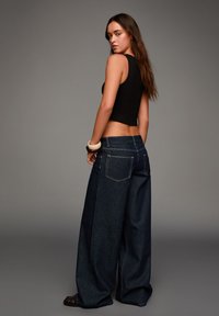 Jeans en denim foncé à jambes larges avec une taille haute et des poches arrière visibles, associés à un crop top noir ajusté, sur fond neutre.