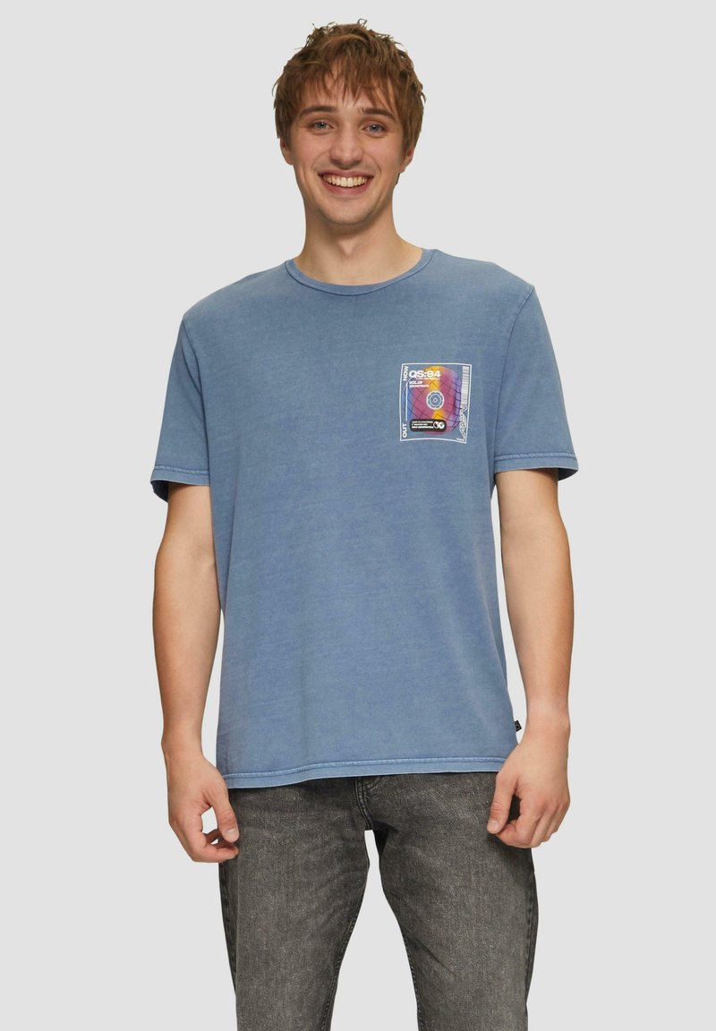 Blauwe katoenen t-shirt met een ronde hals, korte mouwen en een voorzak met een kleurrijk grafisch ontwerp. Gecombineerd met grijze jeans.