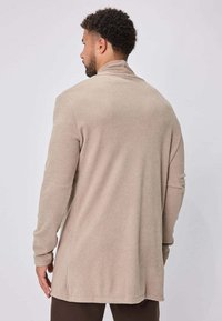 Cardigan à manches longues beige en tissu texturé avec une coupe décontractée, doté de fentes latérales et d'un col montant, conçu dans un style sans coutures.