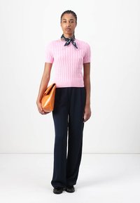 Pull en tricot rose à manches courtes, pantalon large bleu marine, grand sac à main orange, et chaussures noires. Foulard noué autour du cou.