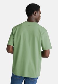 Zelené bavlněné tričko s krátkým rukávem, volný střih a malé vyšívané logo na zadní straně. Bezšvový límec a měkká textura.