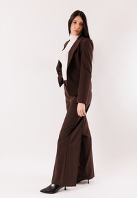 Abito marrone su misura con blazer aderente e pantaloni a gamba larga. Indossato con dolcevita bianco e tacchi neri con punta a mandorla. Materiale liscio.