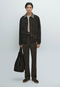 Giacca in suede marrone con un colletto a contrasto, due tasche frontali, abbinata a pantaloni coordinati e una borsa a spalla scura. Texture liscia nel complesso.