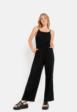 Schwarzer Jumpsuit mit dünnen Trägern, gerafftem Taillenbereich, weitem Bein und seitlichen Taschen. Das Model trägt klobige schwarze Sandalen mit Plateau-Sohle.