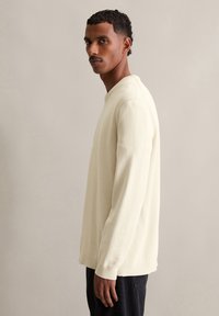 Haut en tricot à manches longues de couleur crème clair avec un col rond et des poignets côtelés, porté avec un pantalon sombre ; il présente un tissu texturé.