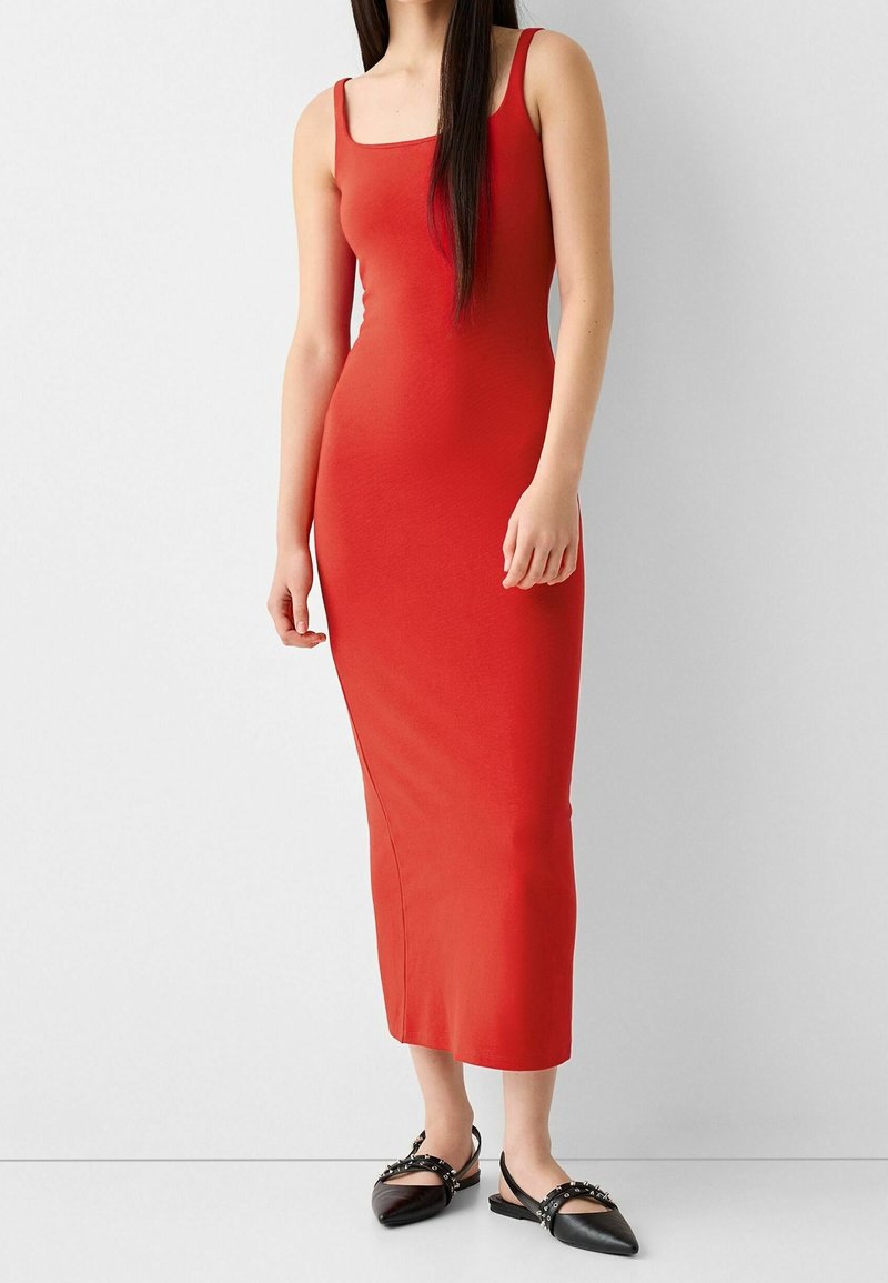 Femme portant une robe midi rouge sans manches et cintrée, associée à des ballerines noires pointues avec des brides cloutées, debout contre un mur blanc.