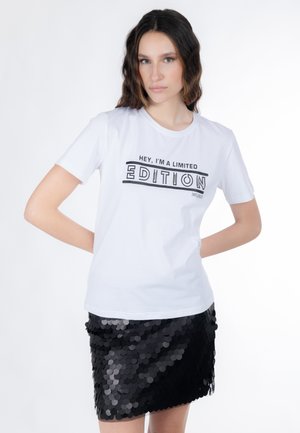 Junge Frau mit welligem Haar trägt ein weißes T-Shirt mit der Aufschrift "Hey, ich bin eine Limited Edition" und einen schwarzen Paillettenrock, steht mit den Händen hinter dem Rücken.