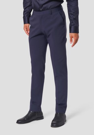Homme portant un pantalon habillé bleu marine foncé ajusté, des bottines en cuir noir et une chemise boutonnée assortie bleu marine foncé, devant un fond uni.