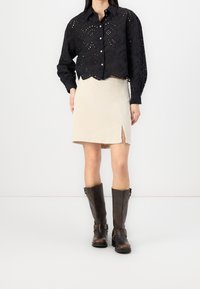 Black embroidered button-up shirt with cut-out patterns, beige slit skirt, and brown knee-high boots with buckles. Minimalist backdrop.