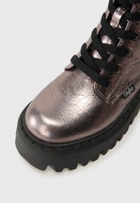 Bota de cuero metálico de color púrpura oscuro con suela de goma negra gruesa y cordones negros, mostrando un primer plano de la puntera y los ojetes.