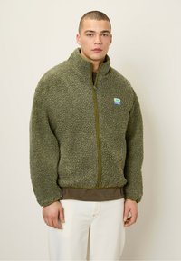 Chaqueta de forro polar verde con un exterior texturizado y esponjoso y una cremallera verde claro. Presenta un pequeño parche colorido de la marca en el pecho.