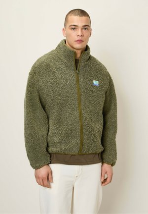 Green fleece zip-up jacket with a textured, fluffy exterior and a light green zipper. Features a small, colorful brand patch on the chest.