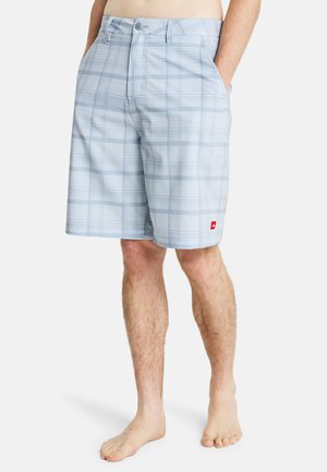 Mężczyzna ubrany w jasnoniebieskie szorty w kratę do kolan, z rękami w kieszeniach, stojący boso na białym tle.