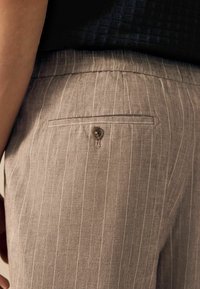 Pantalones de tela a rayas de color marrón claro que cuentan con un bolsillo trasero con cierre de botón. La textura es suave y tiene un corte ajustado.