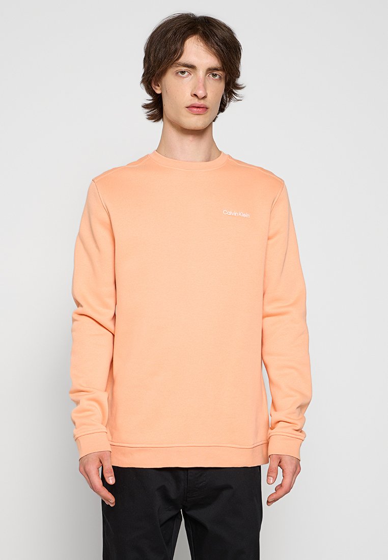 Sweatshirt couleur pêche en coton doux, avec un col rond, des manches longues et un petit logo blanc sur la poitrine.