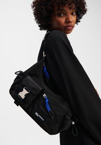 Black fabric shoulder bag with silver hardware, front zip pocket, blue accents, and adjustable strap. Textured surface with a sporty design.