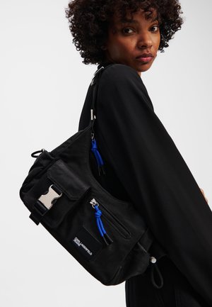 Black fabric shoulder bag with silver hardware, front zip pocket, blue accents, and adjustable strap. Textured surface with a sporty design.
