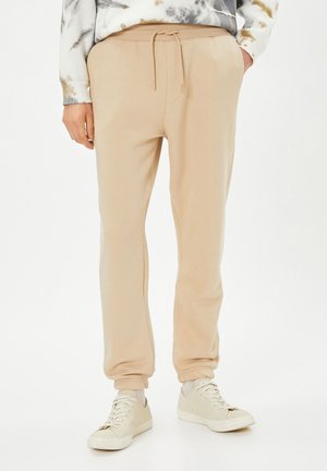 Beige Jogginghosen mit elastischem Bund und Kordelzug. Weiches Material mit lockerer Passform, ausgestattet mit gerafften Säumen und Seitentaschen.