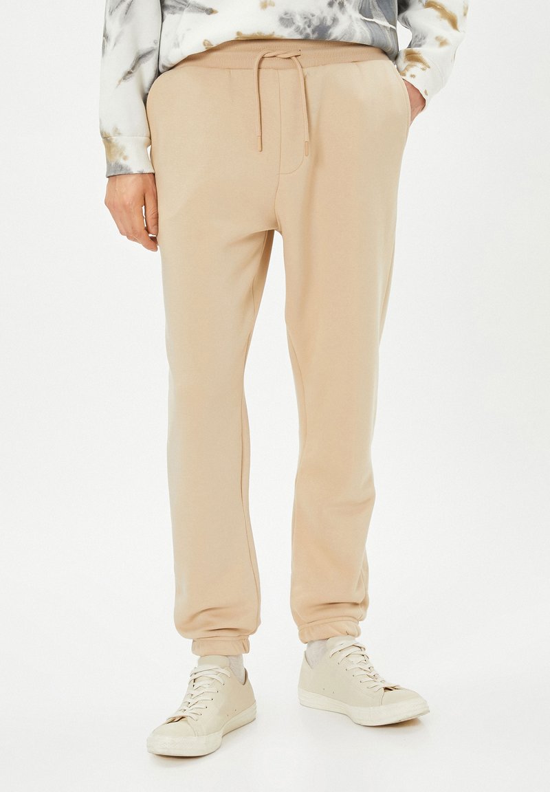Pantalones jogger beiges con cintura elástica y cordón. Tejido suave con un corte relajado, que presenta puños en los tobillos y bolsillos laterales.