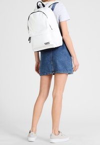 Biały plecak z czarnymi akcentami, z kieszenią z przodu, regulowanymi paskami i logo "TOMMY HILFIGER" na dole. Noszony na sukience dżinsowej.