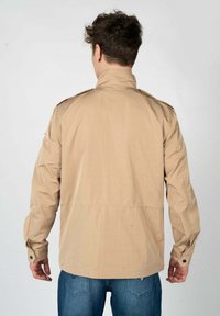 Giacca leggera beige con colletto alto, due epaulette sulle spalle, polsi con bottoni e una texture liscia. Indossata con jeans blu.