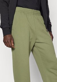 Πράσινο παντελόνι φόρμας με ελαστική μέση, πλαϊνές τσέπες και ελαφρώς υφασμένη επιφάνεια. Συνδυάζεται με μαύρο τζάκετ με φερμουάρ.