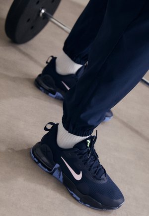 Persoon die zwarte en witte Nike-sneakers en joggings draagt, staand bij een halter met gewichten op de vloer van een sportschool.