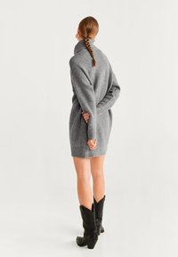 Robe en tricot grise à col roulé, dotée de manches longues. Portée avec des bottes de cowboy noires. Texture lisse, coupe décontractée.