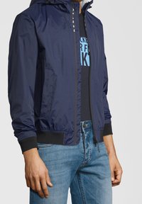 Uomo che indossa una giacca zip navy blu sopra una maglietta nera con grafiche e jeans azzurri chiari, in piedi con le mani rilassate lungo i fianchi.