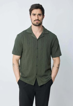 Chemise à manches courtes, boutonnée, de couleur vert foncé, avec un motif géométrique et un col évasé. Portée avec un pantalon noir.