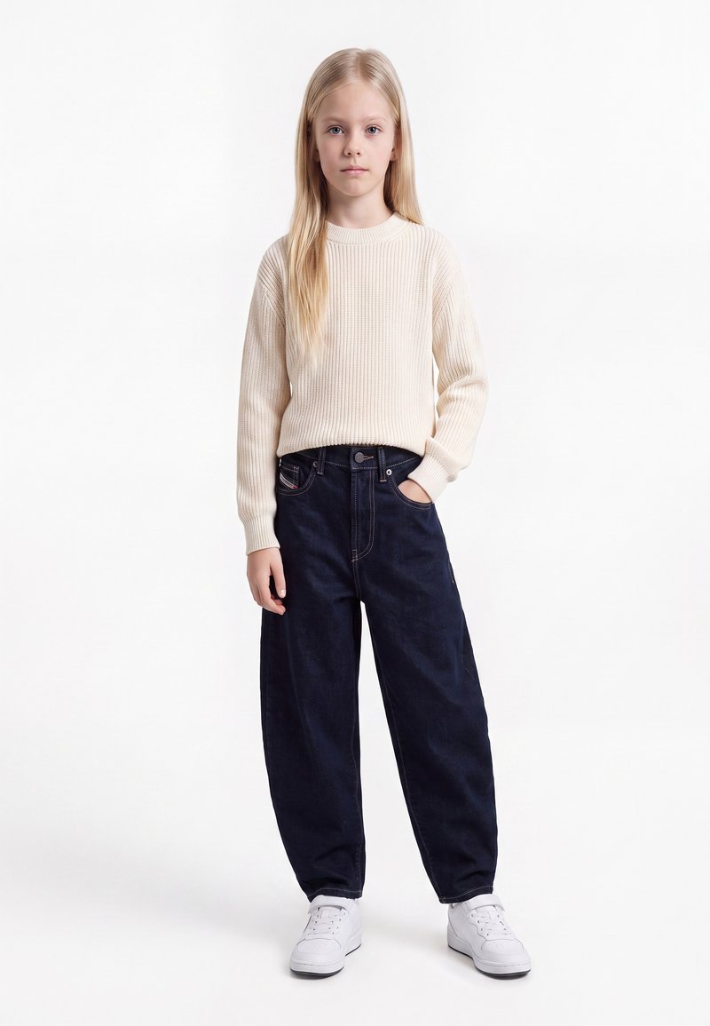 Fille blonde portant un pull en maille crème, un jean bleu foncé ample et des baskets blanches, une main dans la poche.