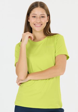 Junge Frau in einem hellgrünen Kurzarmshirt, die lächelt, mit einer Hand am Kinn und dem anderen Arm verschränkt.