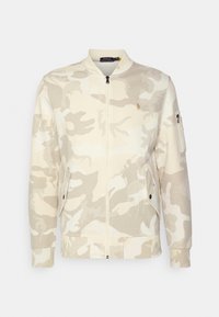 Giacca mimetica beige con collo alto, cerniera frontale, due tasche laterali e una tasca con cerniera sulla manica. Presenta un logo sul petto.