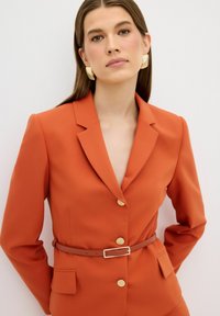 Oranje gemaakt blazer met een notch lapel en gouden knopen. Bevat een slanke bruine riem en zijzakken. Gladde stoftextuur.