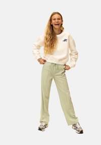 Witte sweatshirt met blauw sterlogo, gecombineerd met lichtgroene broek met elastische taille en grijze sneakers. Model staat met de handen in de zij.