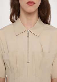 Beige ing rövid gallérral és elöl zipzárral, két nagy zsebbel és függőleges redőzéssel a textúráért. Sima anyagfelület.