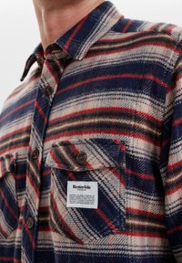 Camisa de franela con un patrón de cuadros en azul, rojo y beige. Tiene un cierre delantero con botones y un bolsillo en el pecho con una etiqueta de la marca.