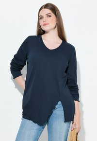 Maglia a maniche lunghe blu navale con scollo a V e dettagli a orlo grezzo, abbinata a jeans azzurri. Il materiale sembra morbido con una vestibilità rilassata.