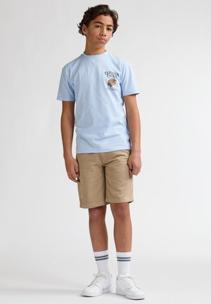 Tricou bleu deschis din bumbac cu grafică taco, asortat cu pantaloni scurți kaki. Pantofi sport albi și șosete scurte în dungi completează ținuta.