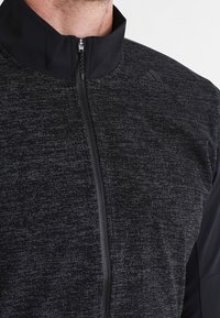 Hombre con chaqueta con cremallera texturizada negra de Adidas, con collar alto y logo visible en la parte superior derecha del pecho.