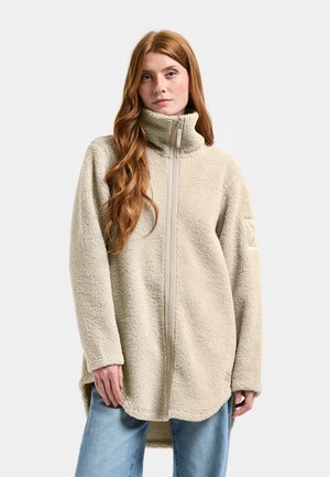 Kobieta o długich rudych włosach, ubrana w beżową polarkę z wysokim kołnierzem i jasnoniebieskie dżinsy, stojąca na tle jednolitego szarego tła.