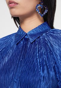 Camicia blu in satin con texture a pieghe, colletto a punta e bottoni; presenta grandi orecchini a forma di cuore con gemme blu.