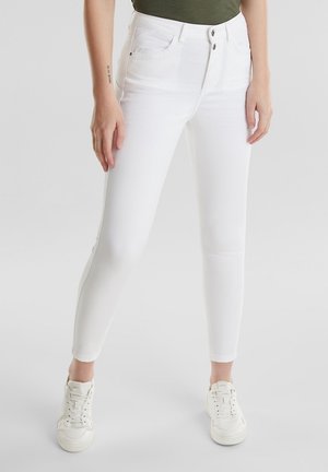 Vaqueros blancos de corte ajustado con cierre de doble botón usados con zapatillas blancas en una persona que está de pie en el interior frente a un fondo liso.