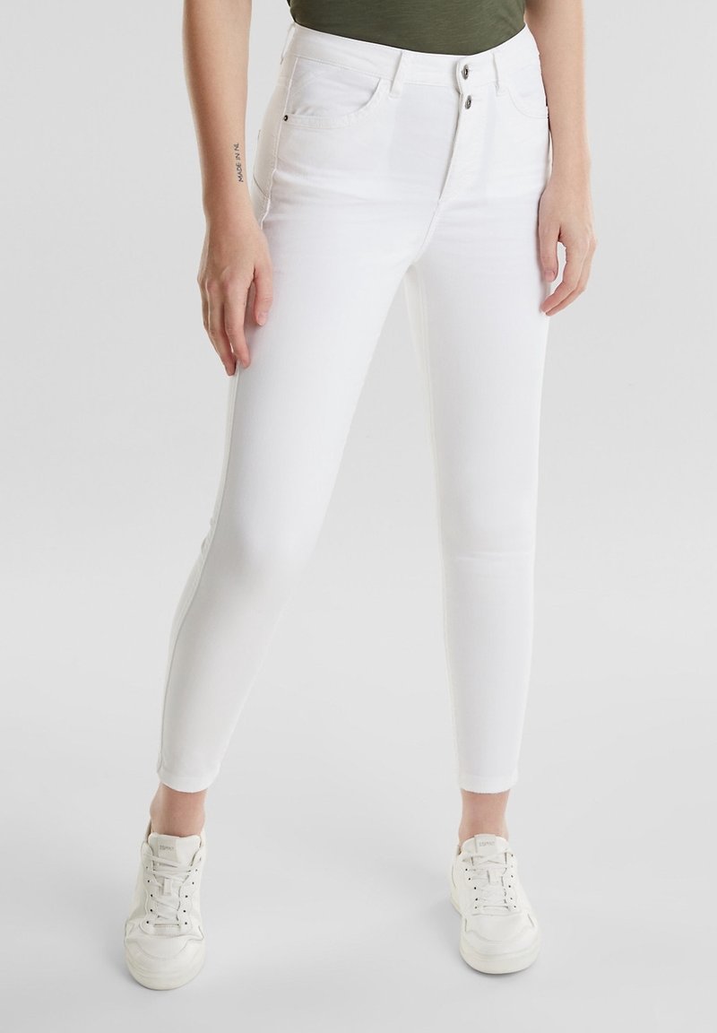 Jean blanc slim-fit avec fermeture double bouton porté avec des baskets blanches sur une personne debout à l'intérieur devant un fond uni.