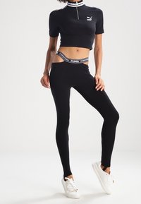 Žena v černém krajkovém topu Puma a legínách s vysokým pasem a detailním výřezem, v bílých teniskách, stojí na jednobarevném pozadí.