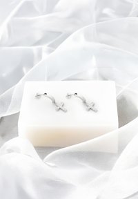 Silverfärgade hoopsörhängen med ett korsdesign, dekorerade med klara rhinestones, placerade på en vit yta mot mjuk, flödande tyg.