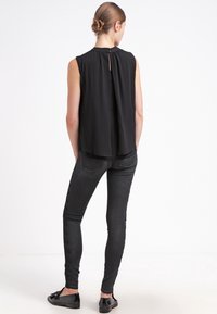 Černý top bez rukávů se zadním detaily ve tvaru klíčové díry, kombinovaný s úzkými černými džíny a plochými botami, zobrazený zezadu.